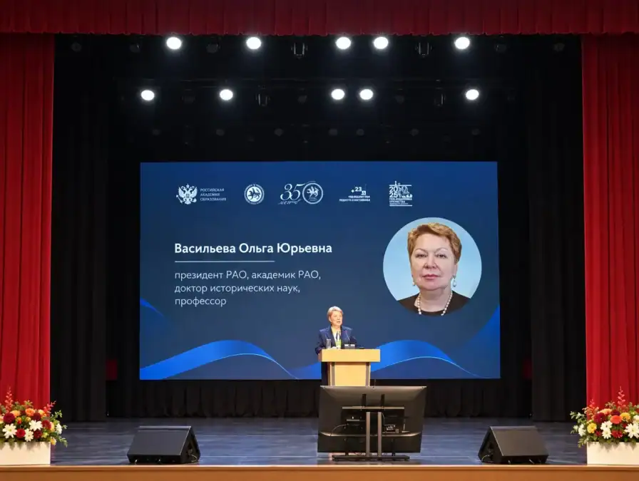 Спикер выступает за трибуной на сцене перед большим информационным экраном.