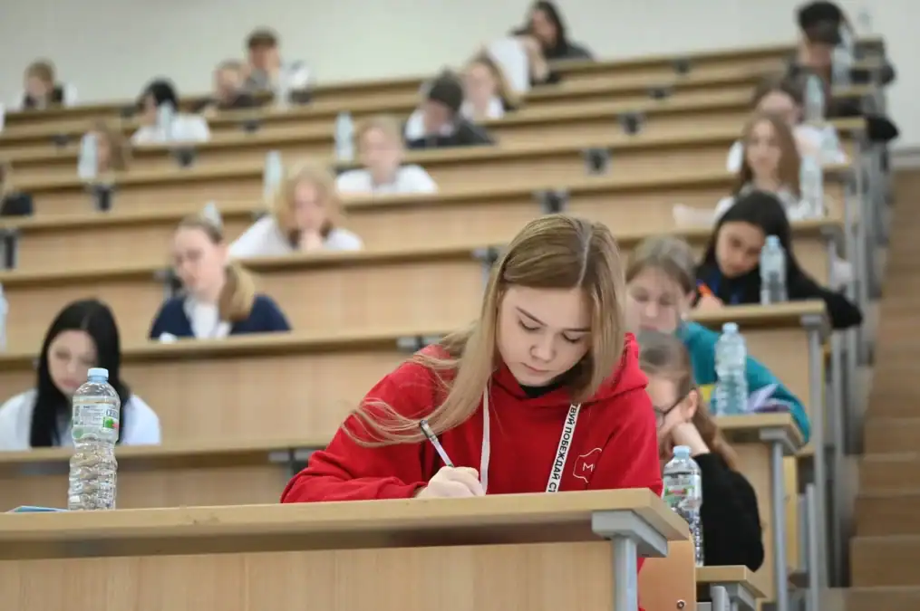 Участница в красном свитере пишет работу в заполненной школьниками аудитории.