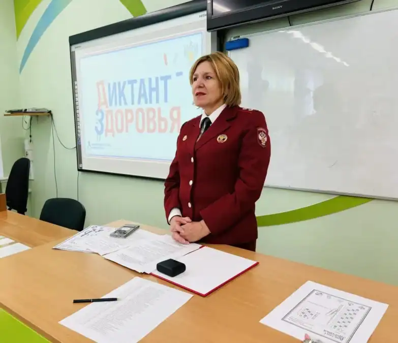 Женщина в форменной одежде стоит у стола перед экраном «Диктант здоровья».