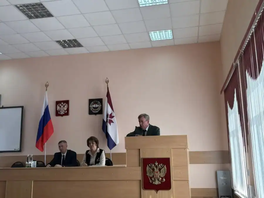 Мужчина выступает с трибуны, рядом за столом сидят двое участников заседания.