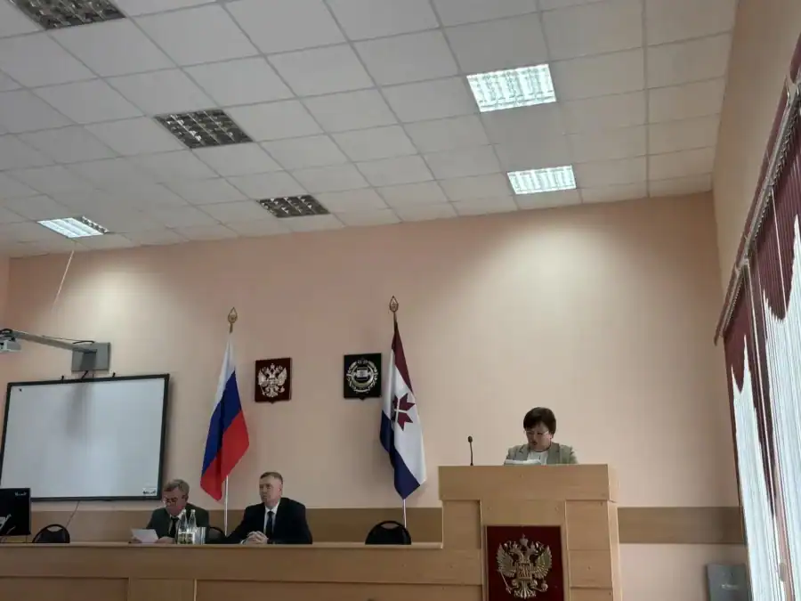 Женщина выступает с трибуны, двое мужчин сидят за столом президиума.