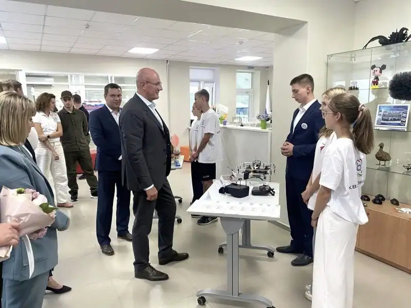 Взрослые и школьники стоят у стола с техническими моделями в учебном помещении.