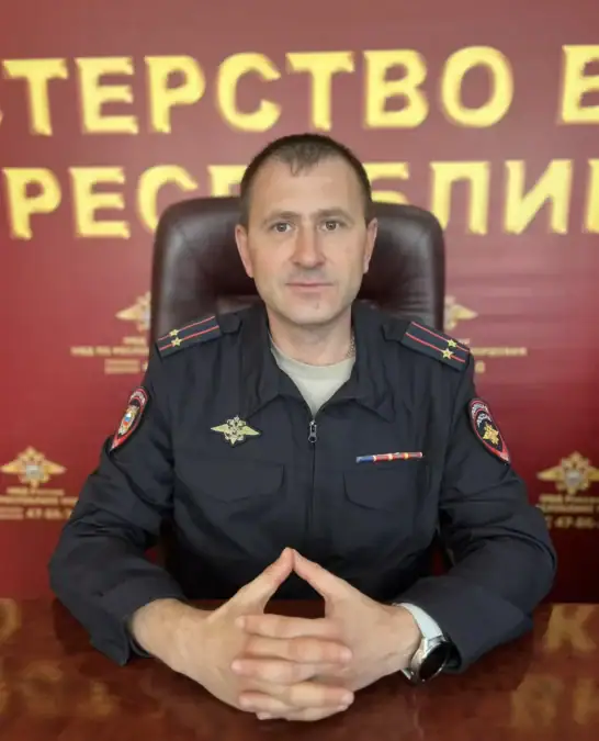 Сотрудник МВД в форме сидит за столом на фоне стены с символикой ведомства.
