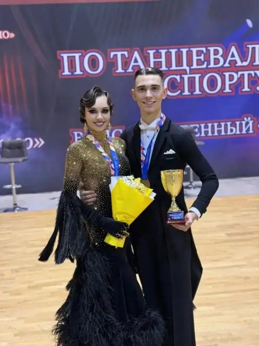 Танцевальная пара с медалями, кубком и букетом стоит на паркете.