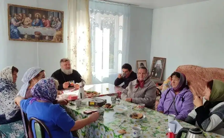 Пожилые женщины и священник сидят за накрытым столом в комнате.