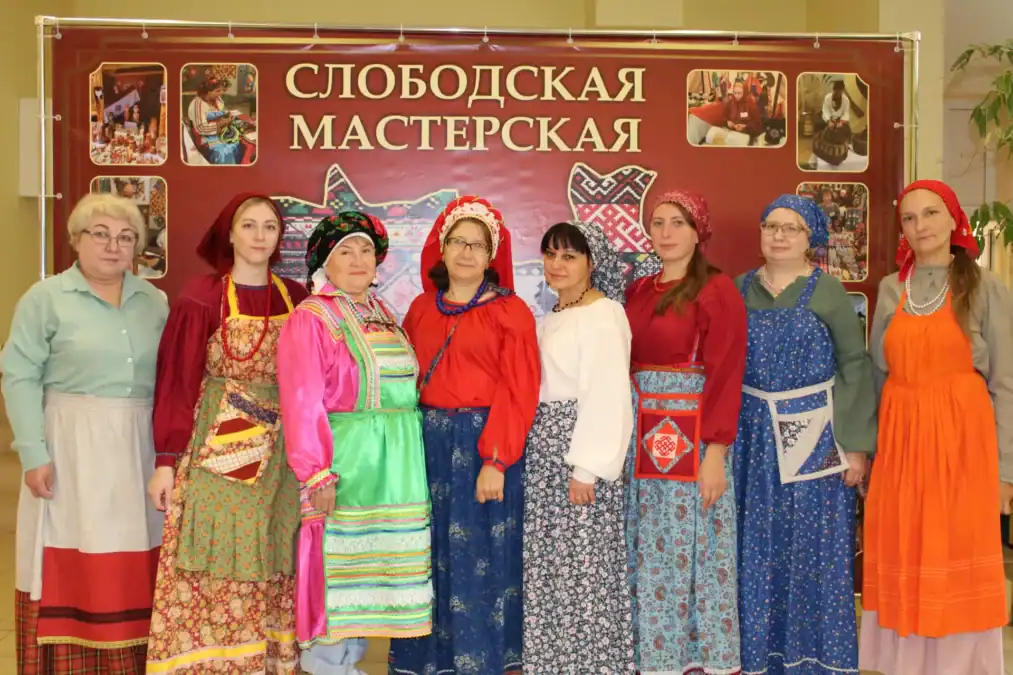 Восемь женщин в народных костюмах стоят перед баннером «Слободская мастерская».