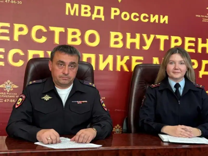 Двое сотрудников полиции сидят за столом перед стендом МВД Мордовии.