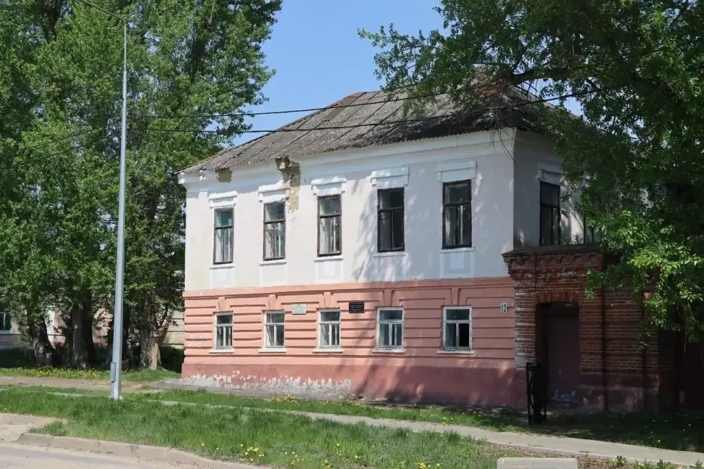 Двухэтажное белое историческое здание с кирпичной пристройкой среди деревьев.