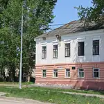 Двухэтажное белое историческое здание с кирпичной пристройкой среди деревьев.