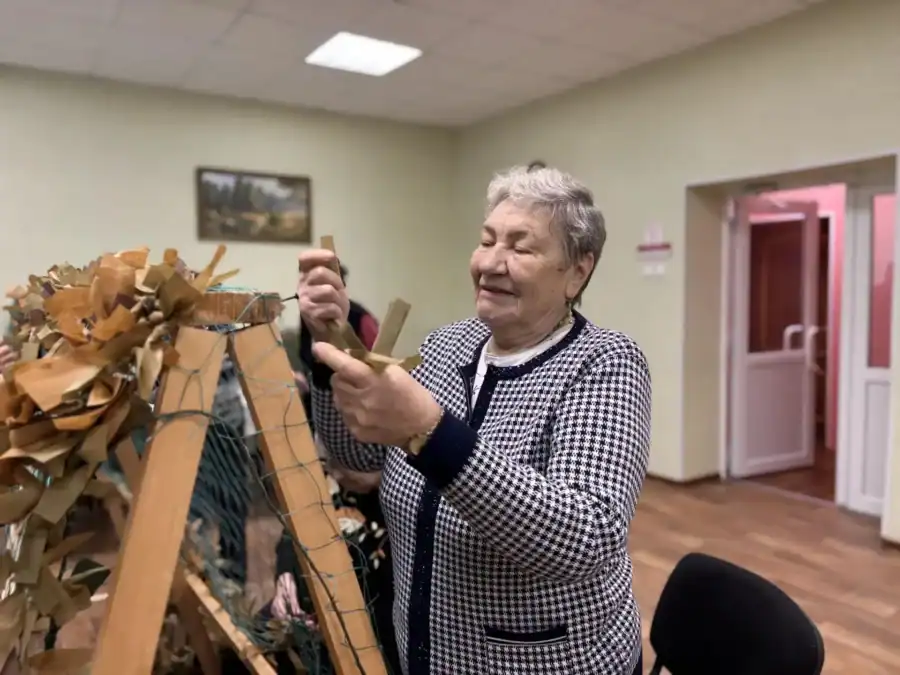 Пожилая женщина работает с плетёной декоративной композицией в помещении.