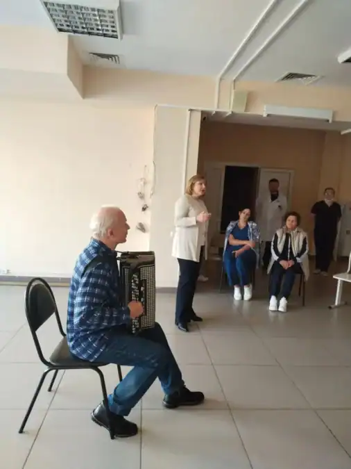 Баянист и женщина выступают перед сидящими слушателями в коридоре.