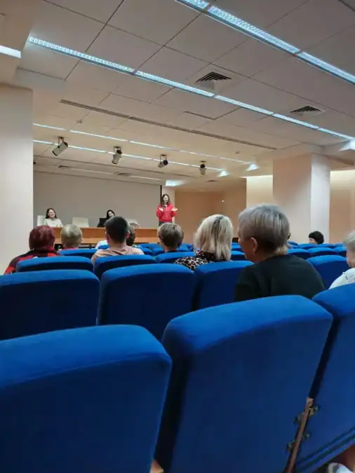 Участники сидят в синем зрительном зале перед выступающей женщиной.