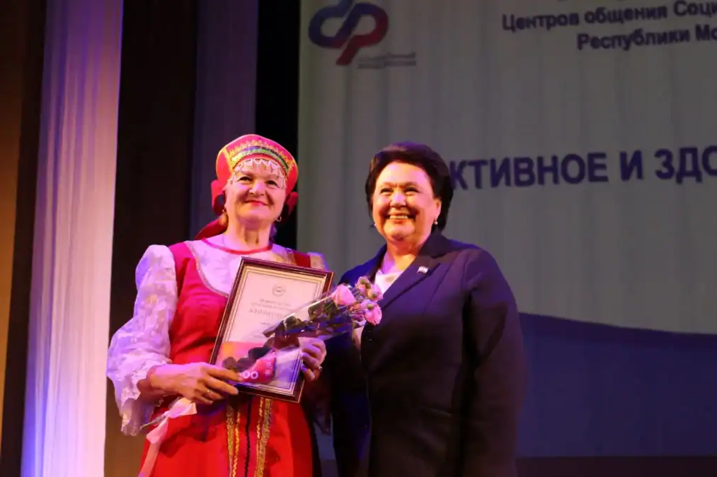 Женщина в народном костюме держит диплом и букет рядом с участницей мероприятия.