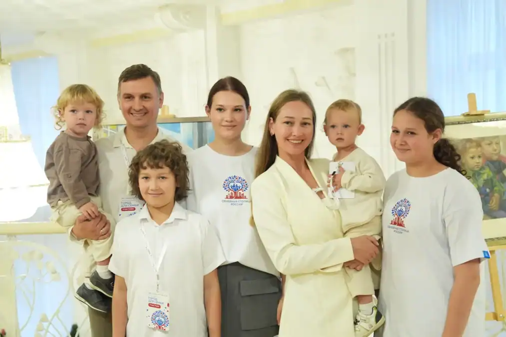 Семья с детьми позирует рядом с участниками форума в светлом зале.