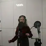 Девушка в наушниках стоит перед микрофоном в звукозаписывающей кабине.