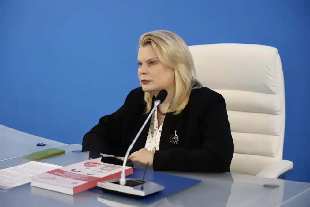 Женщина выступает за столом с микрофоном и печатными материалами.