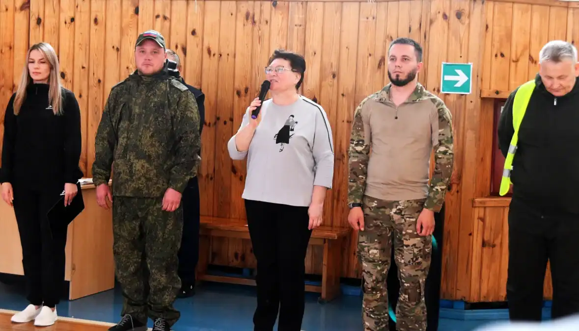 Женщина с микрофоном стоит рядом с участниками в спортивном зале.