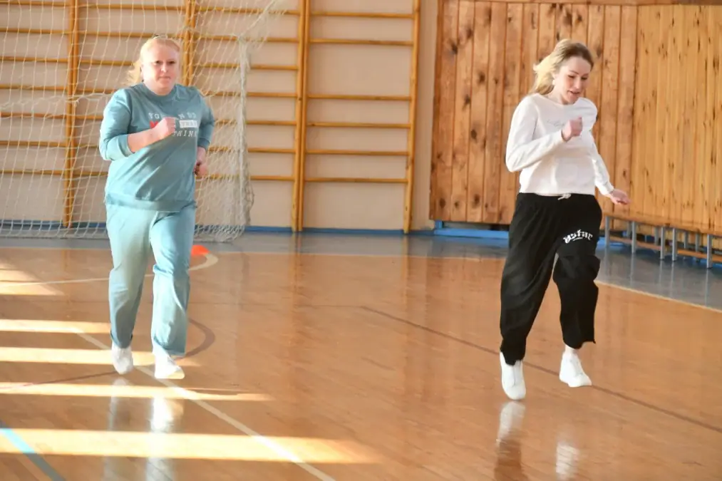 Две женщины бегут по спортивному залу с деревянными стенами и сеткой.