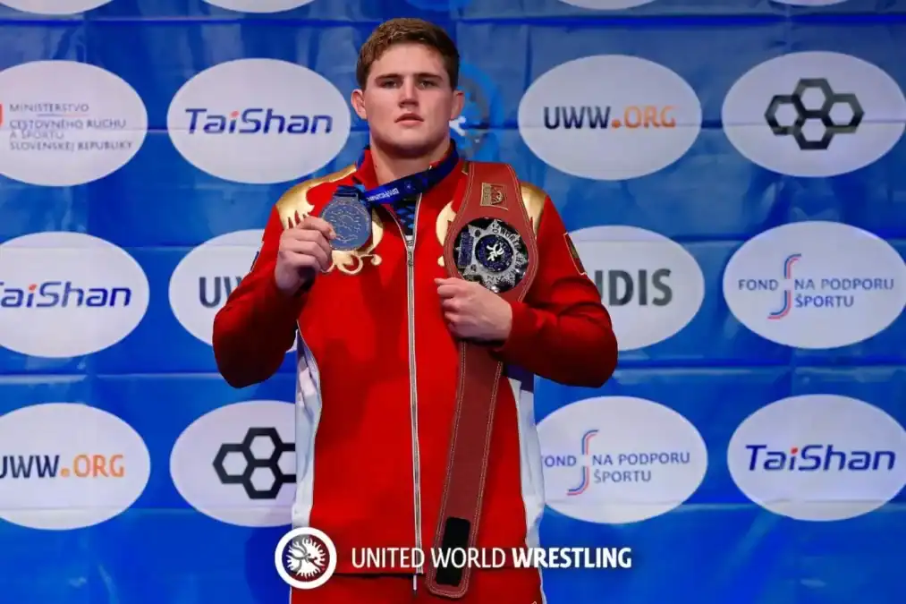 Спортсмен в красной спортивной куртке показывает медаль и чемпионский пояс.