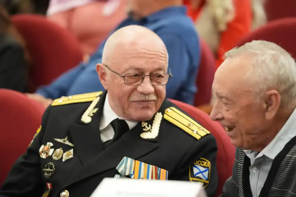 Ветеран в форме с медалями беседует с мужчиной в зрительном зале.