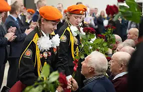 Курсантки МЧС вручают красные гвоздики сидящим ветеранам.