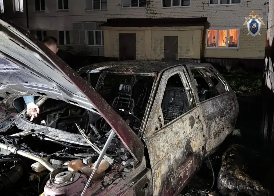 Сильно выгоревший автомобиль с открытым капотом стоит у жилого дома ночью.