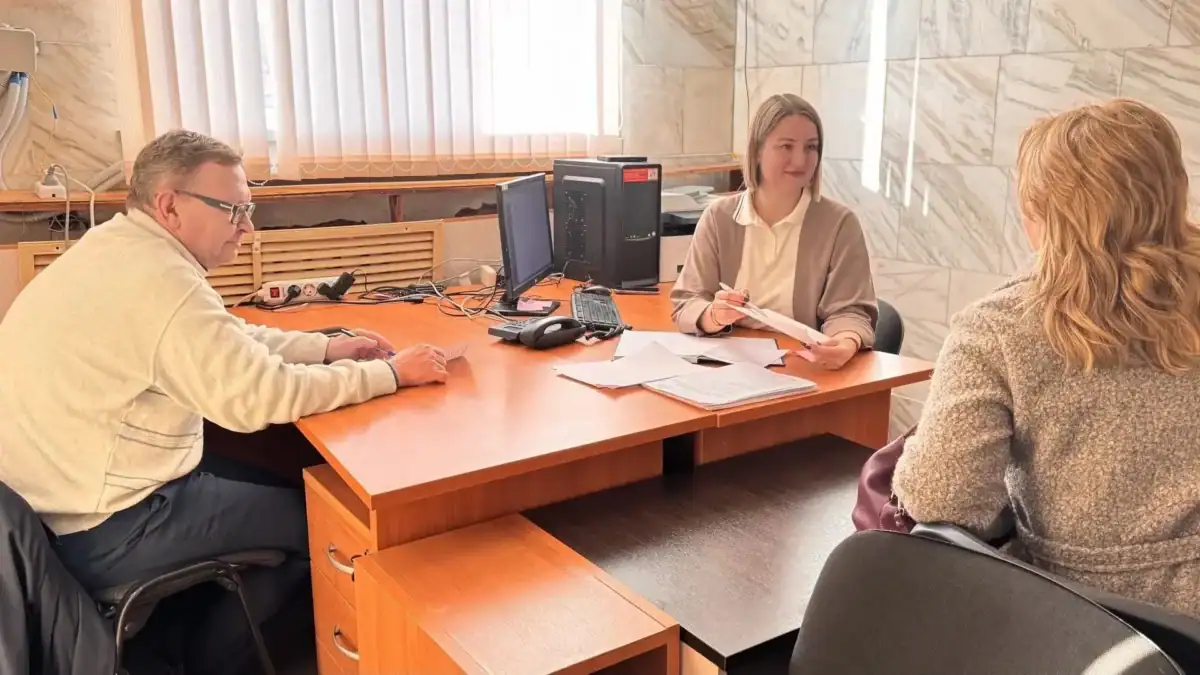 Сотрудница за столом беседует с двумя посетителями и просматривает документы.