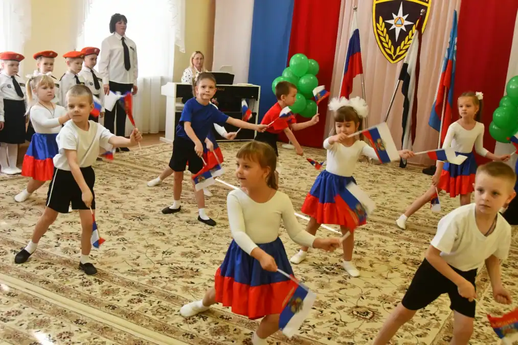 Дети исполняют танец с российскими флагами в украшенном зале.