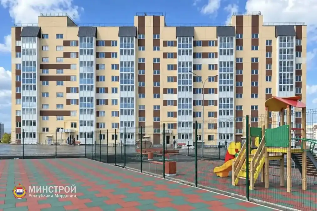 Новый многоэтажный жилой дом за ограждённой детской площадкой.