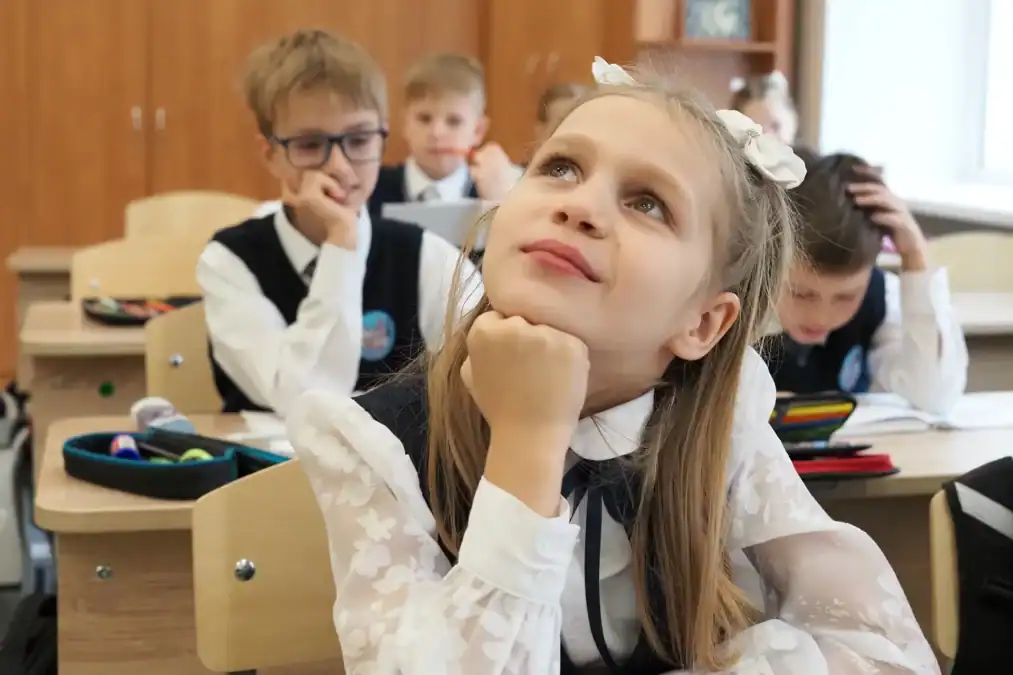 Школьница сидит за партой, положив подбородок на руку.