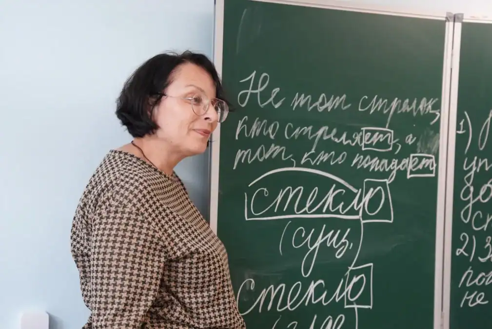 Учительница стоит у доски с написанным русским текстом.
