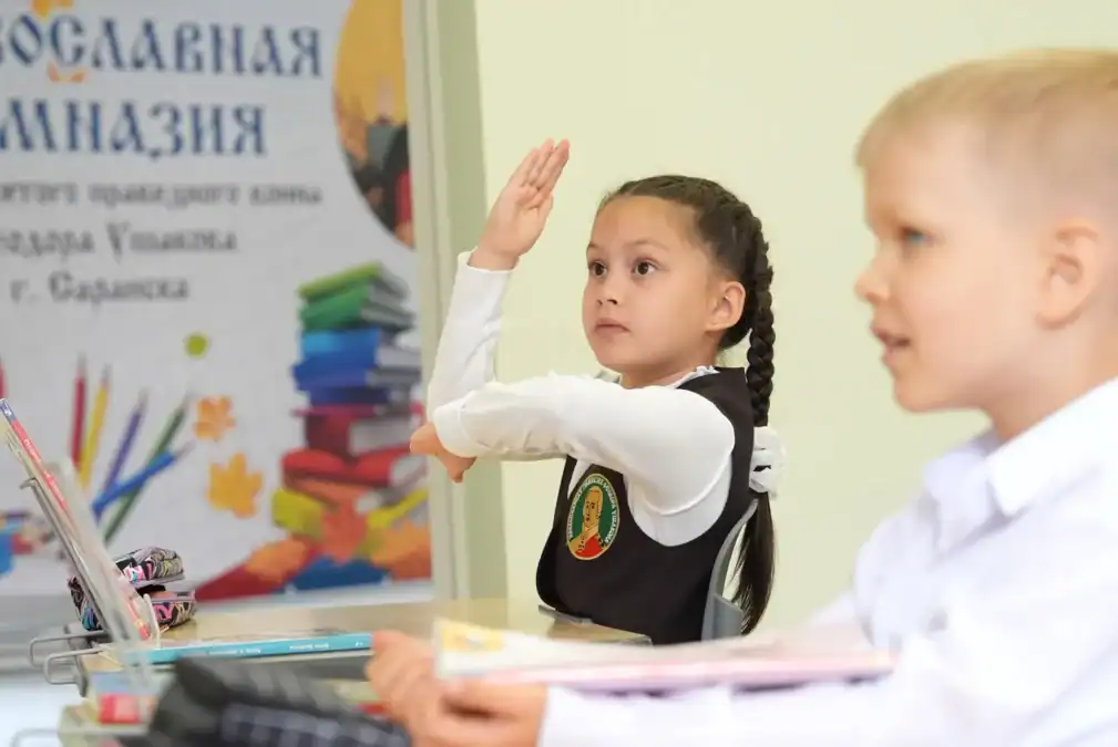 Школьница поднимает руку во время занятия за партой.