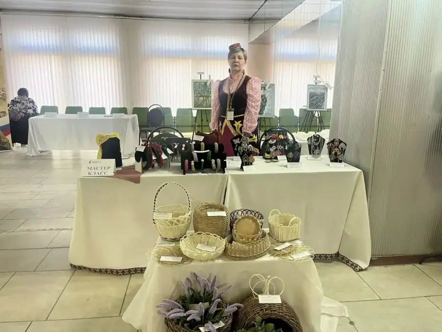Женщина стоит за столом с украшениями, куклами и плетёными корзинами.