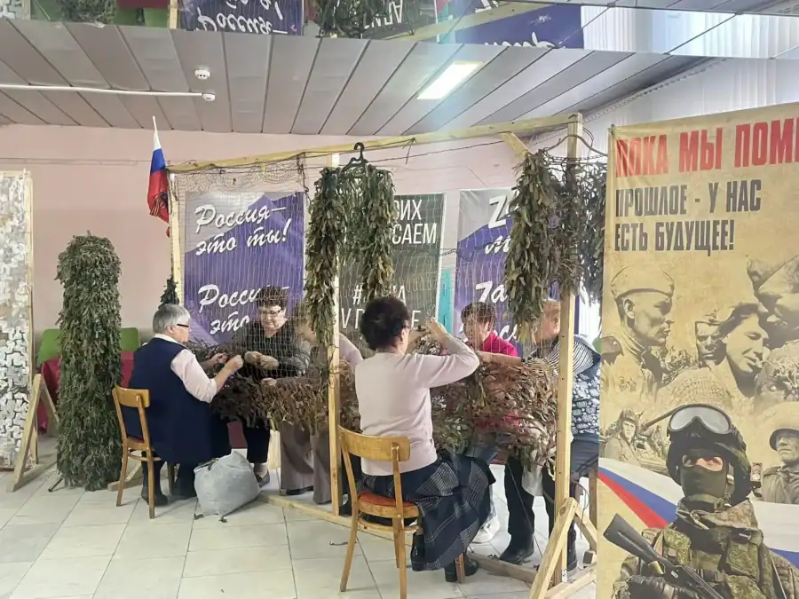 Посетители за столом плетут маскировочную сеть рядом с тематическим стендом.
