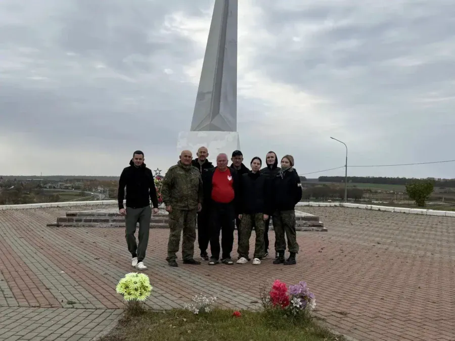 Группа людей стоит у мемориала, перед которым лежат букеты цветов.