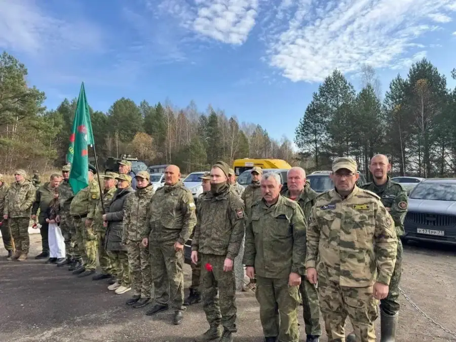 Поисковики в камуфляже стоят строем рядом с флагом и автомобилями.