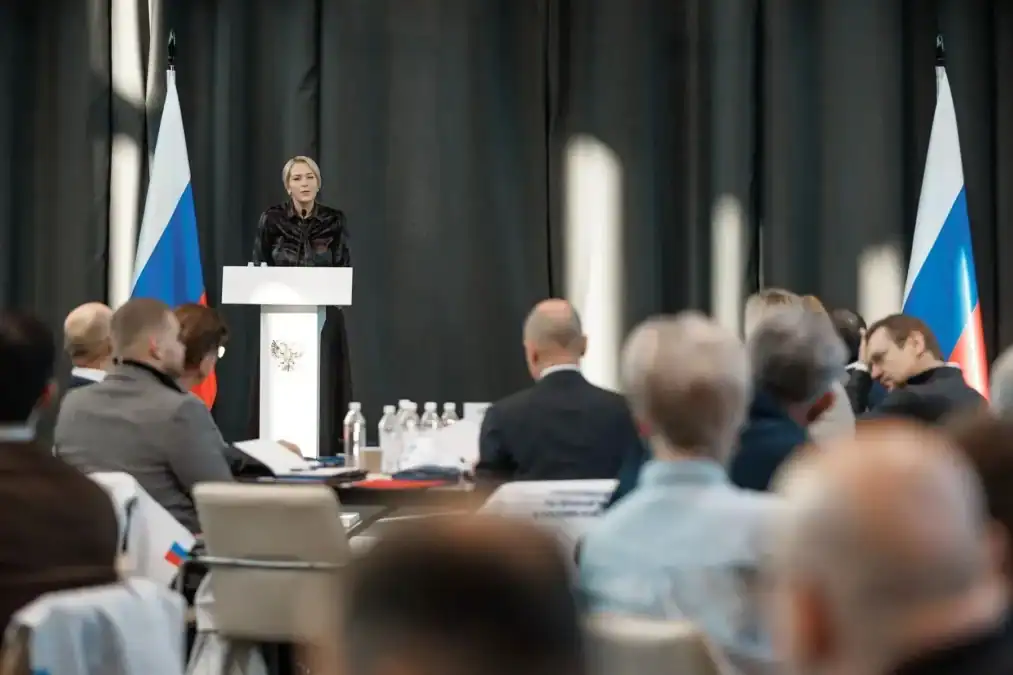 Женщина выступает с трибуны между российскими флагами.
