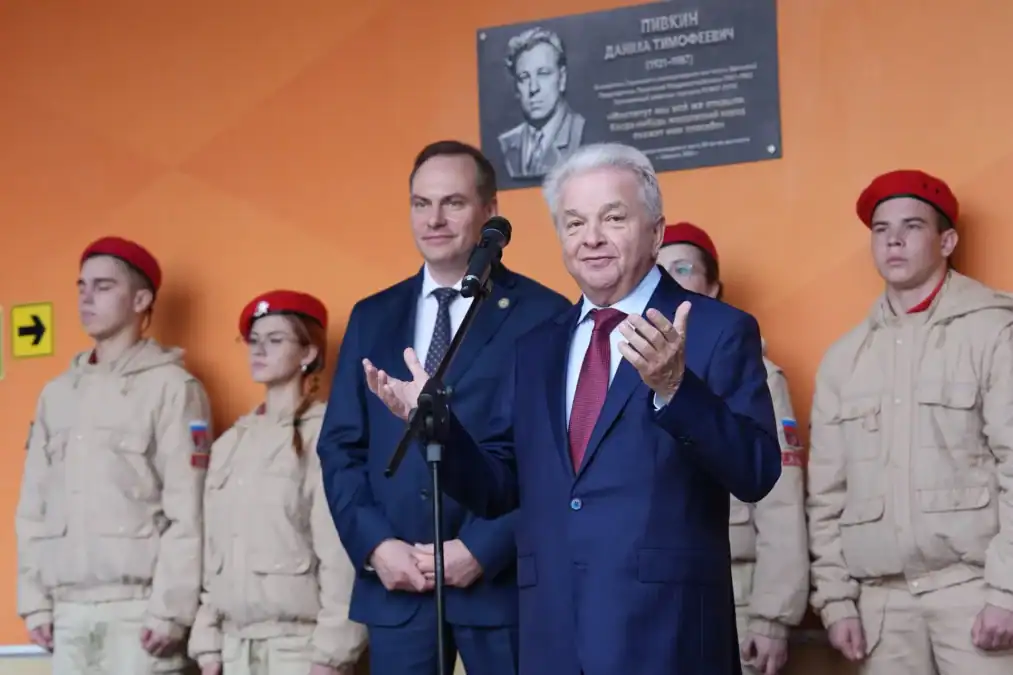 Мужчина выступает у микрофона на фоне мемориальной доски и юнармейцев.