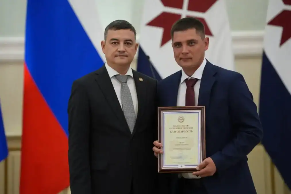 Двое мужчин позируют с дипломом на фоне российских и региональных флагов.