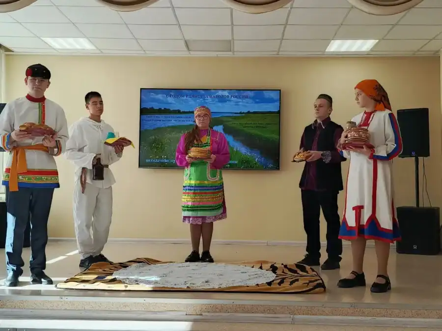 Участники в народных костюмах стоят на сцене вокруг женщины с букетом.