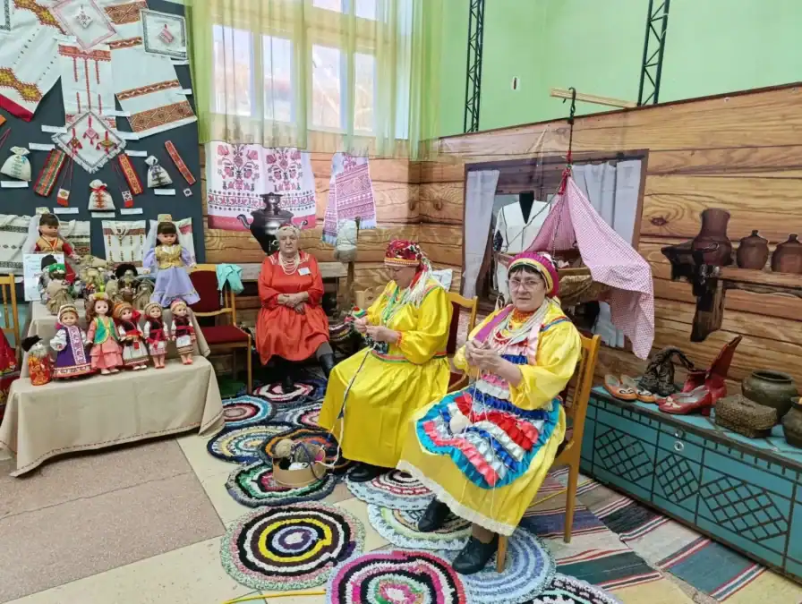 Участники в народных костюмах сидят среди кукол, тканей и предметов ремесла.