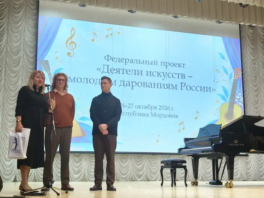 Три человека стоят у микрофона на сцене рядом с роялем и экраном проекта.
