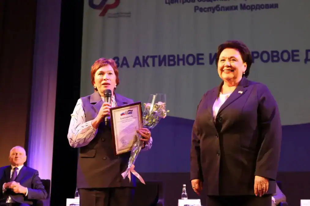 Женщина с дипломом и букетом стоит на сцене рядом с участницей мероприятия.