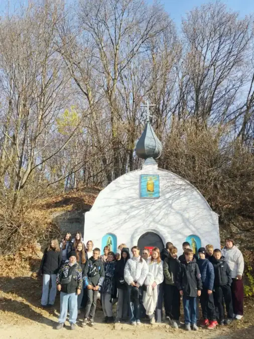 Группа школьников стоит у входа в небольшую белую часовню среди деревьев.