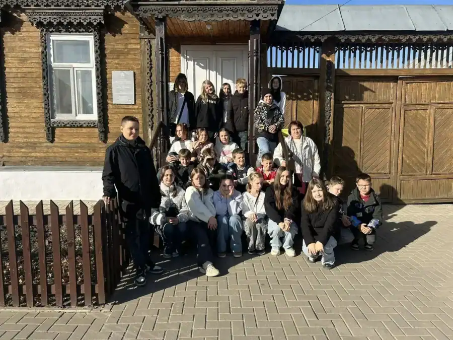 Школьники позируют у деревянного дома с резными наличниками и воротами.