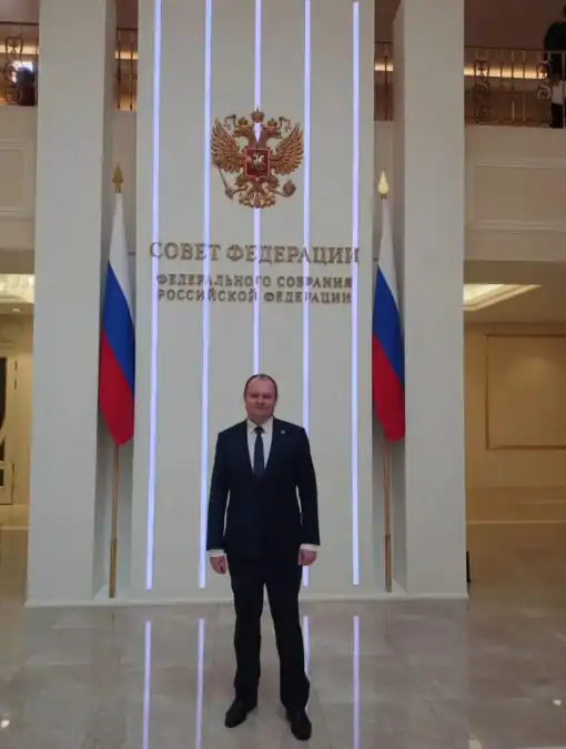 Мужчина в костюме стоит у стены с гербом и надписью Совета Федерации.