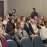 Участники мероприятия сидят в зале, несколько человек стоят у стены.