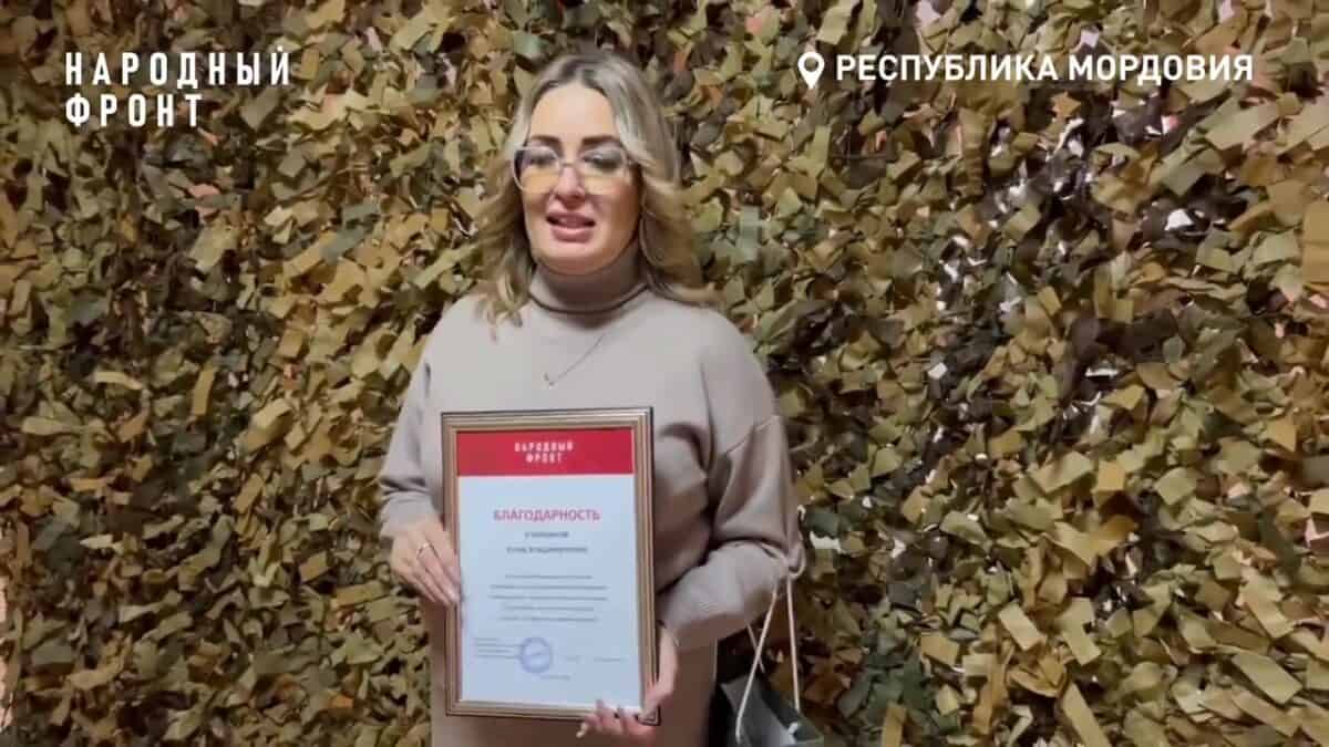 Женщина держит в руках благодарственное письмо на фоне маскировочной сети.