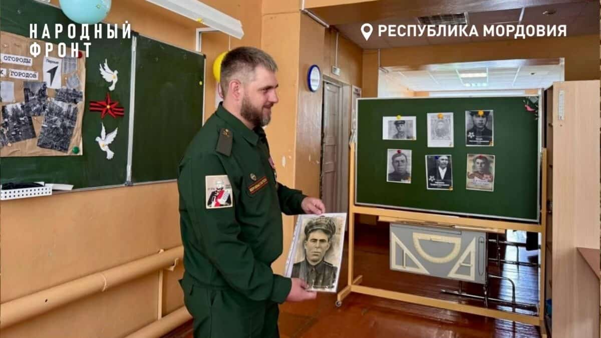Мужчина в военной форме держит портрет в школьном коридоре у стендов.