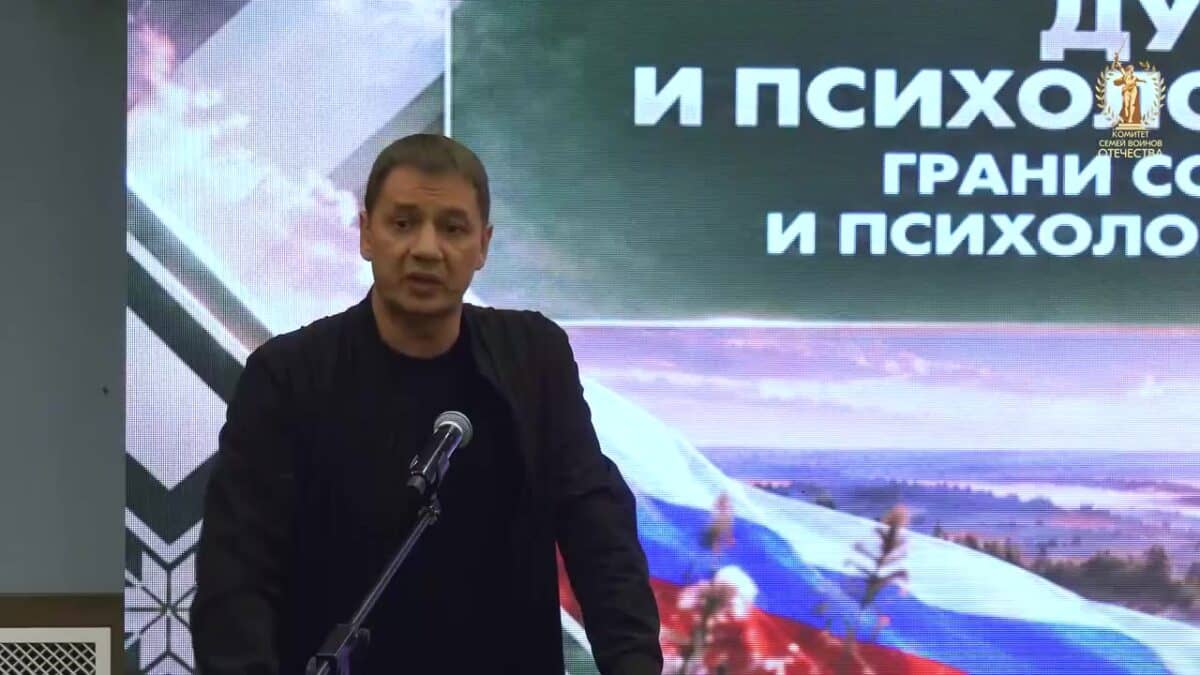 Мужчина выступает у микрофона перед экраном с изображением российского флага.